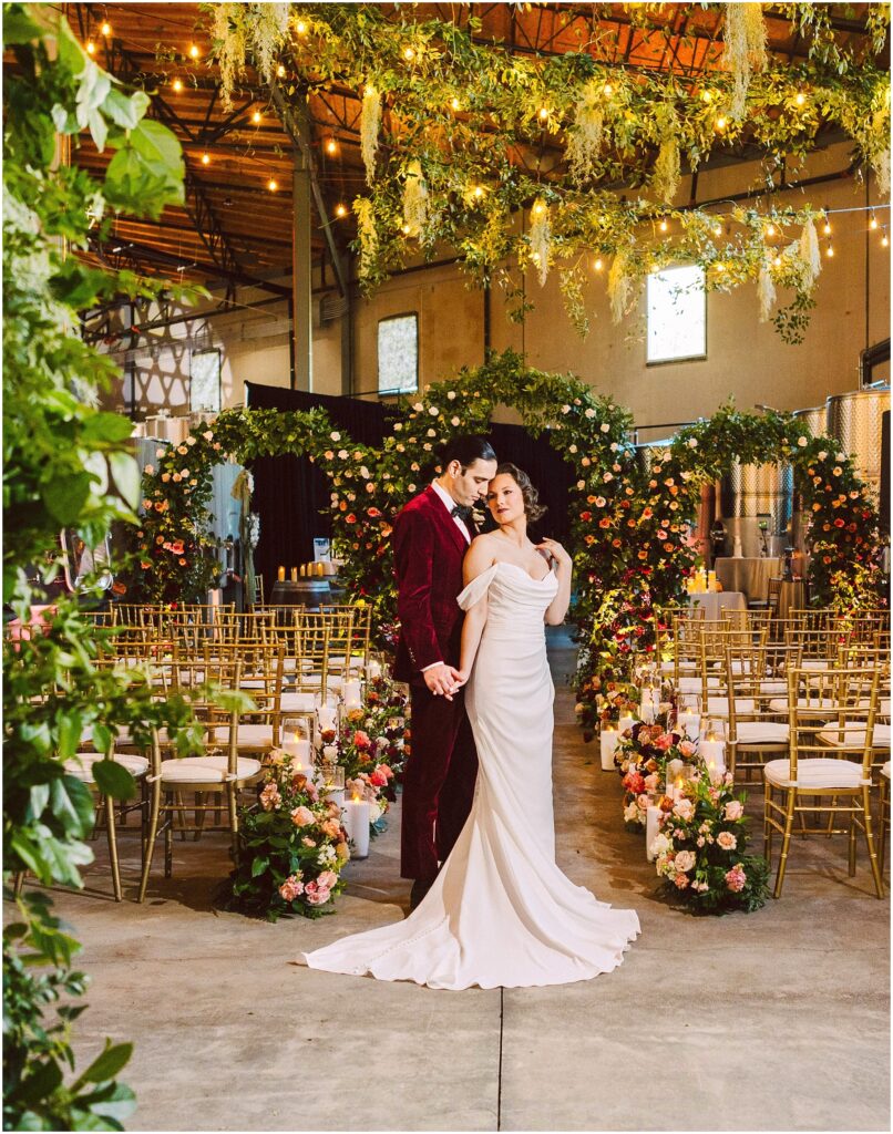 Snohomish Wedding Photographer GSquared Weddings A couple stands close together in a wedding venue, surrounded by gold chairs and vibrant floral arrangements. The bride wears an elegant white gown, and the groom is in a red velvet suit. Greenery and string lights are suspended above them. Serving Seattle, Snohomish and Orlando
