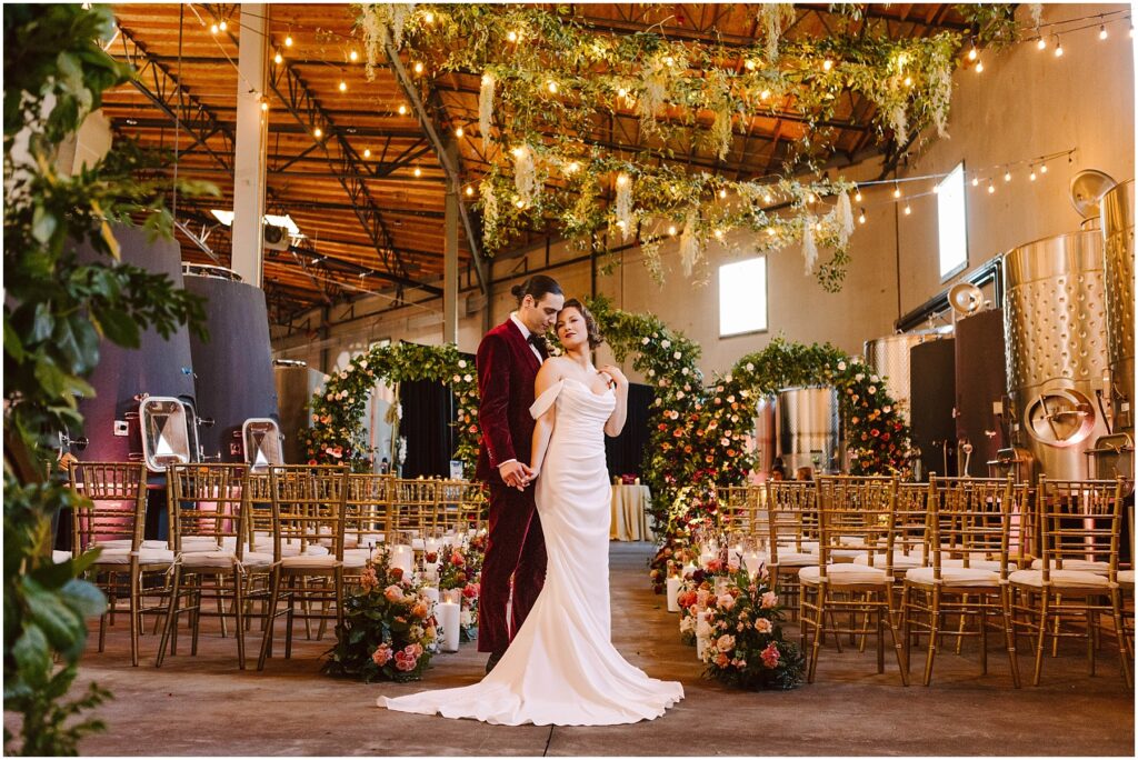 Snohomish Wedding Photographer GSquared Weddings A bride and groom stand in an elegantly decorated industrial venue. The groom wears a burgundy suit, and the bride wears a white dress. There are floral arrangements and chairs arranged for an event. String lights and greenery hang overhead. Serving Seattle, Snohomish and Orlando