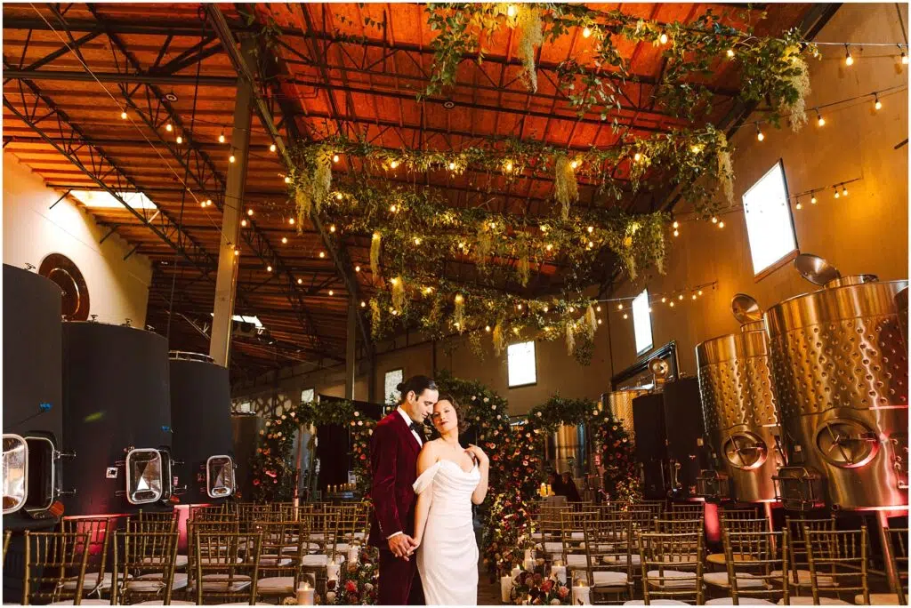 Snohomish Wedding Photographer GSquared Weddings A bride in a white dress and a groom in a burgundy suit stand close in a warmly lit industrial venue decorated with greenery and string lights. Rows of chairs and large metal tanks are visible in the background. Serving Seattle, Snohomish and Orlando