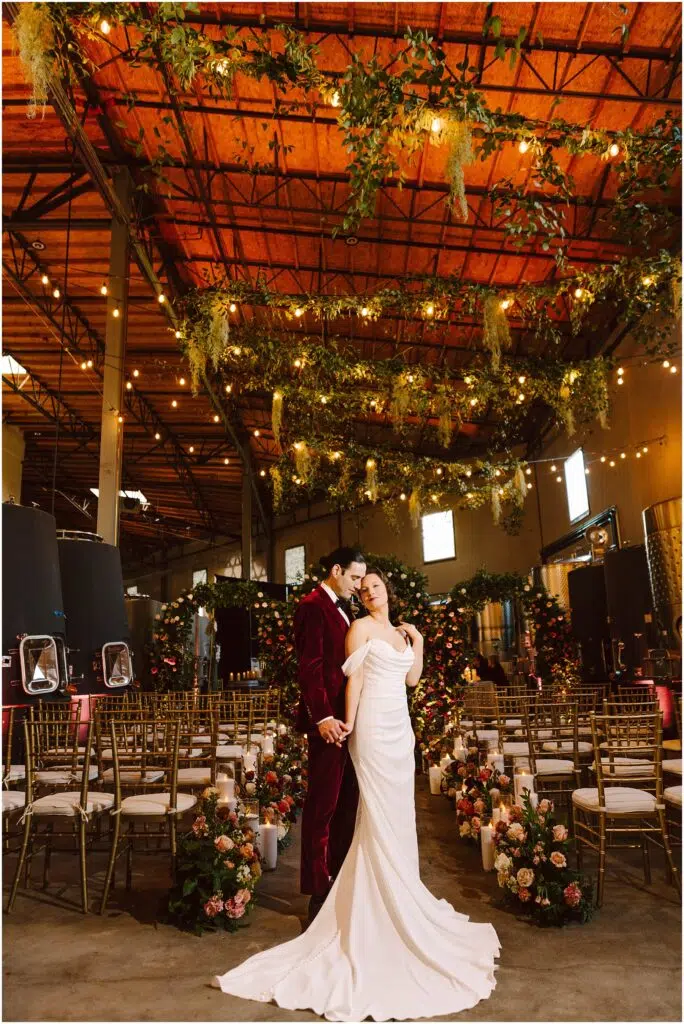 Snohomish Wedding Photographer GSquared Weddings A couple poses in a decorated indoor wedding venue. The bride wears a white gown, and the groom dons a red suit. The space is adorned with hanging greenery, string lights, and abundant floral arrangements, with rows of chairs for guests. Serving Seattle, Snohomish and Orlando