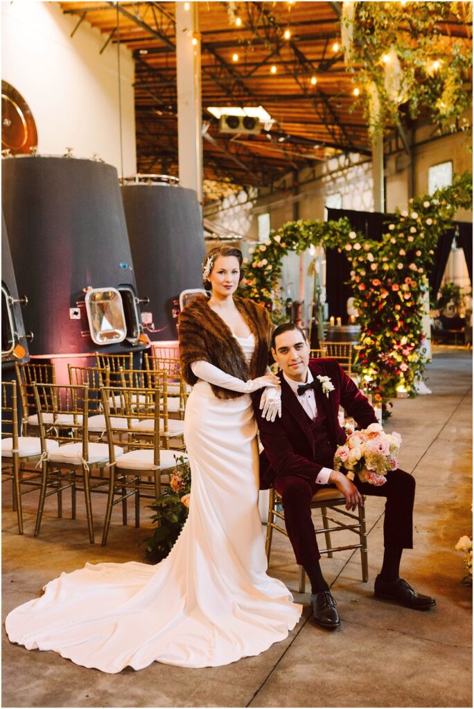 Snohomish Wedding Photographer GSquared Weddings A couple in vintage attire poses in a beautifully decorated venue. The woman in a white gown and fur stole stands beside the seated man in a burgundy suit. Floral arrangements and rows of chairs are visible in the background. Serving Seattle, Snohomish and Orlando