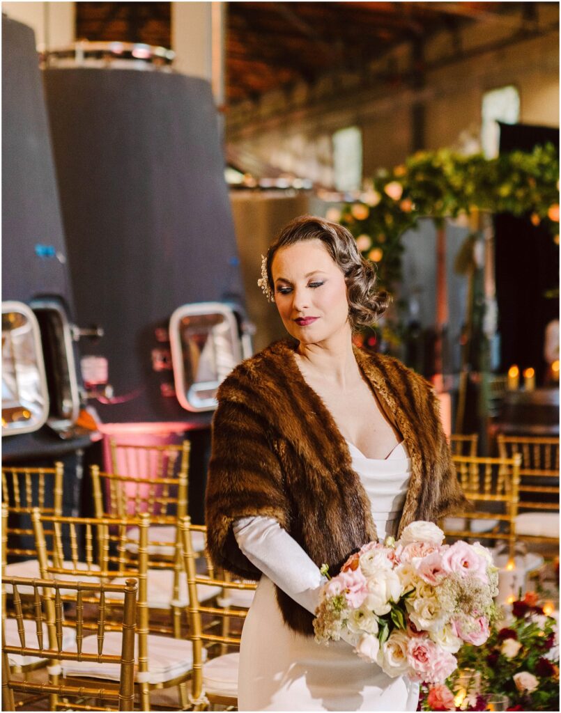 Snohomish Wedding Photographer GSquared Weddings A woman in a fur shawl holds a bouquet of pink and white flowers. She stands in a venue with rows of gold chairs, and large wine tanks are visible in the background. The setting suggests a rustic and elegant event atmosphere. Serving Seattle, Snohomish and Orlando