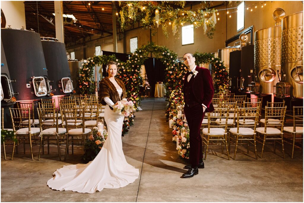 Snohomish Wedding Photographer GSquared Weddings A bride and groom stand in the center of an aisle adorned with flowers in an industrial-style venue. She wears a white gown and fur wrap, holding a bouquet. He wears a burgundy suit. Rows of gold chairs and string lights enhance the setting. Serving Seattle, Snohomish and Orlando