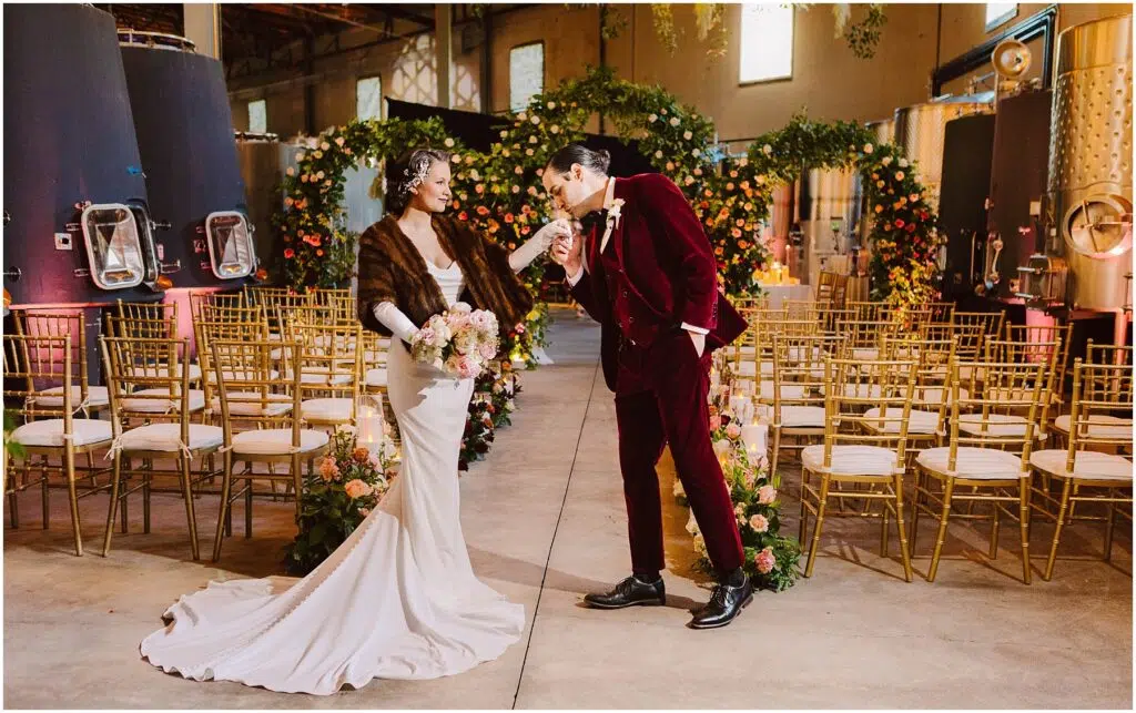 Snohomish Wedding Photographer GSquared Weddings A couple stands in an elegant setting with gold chairs and colorful floral arrangements. The woman, in a white gown and brown shawl, holds a bouquet, while the man, in a burgundy suit, bows gently. The venue is adorned with decorative arches. Serving Seattle, Snohomish and Orlando