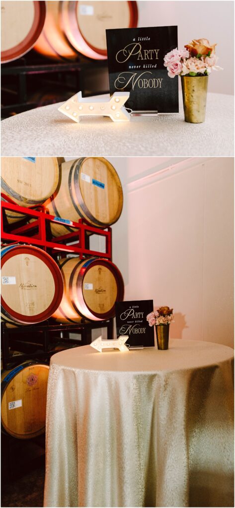Snohomish Wedding Photographer GSquared Weddings A small round table with a gold tablecloth holds a lit arrow sign, a black sign with cursive text, and a vase with pink and peach flowers. Behind the table are stacked wooden barrels. Serving Seattle, Snohomish and Orlando