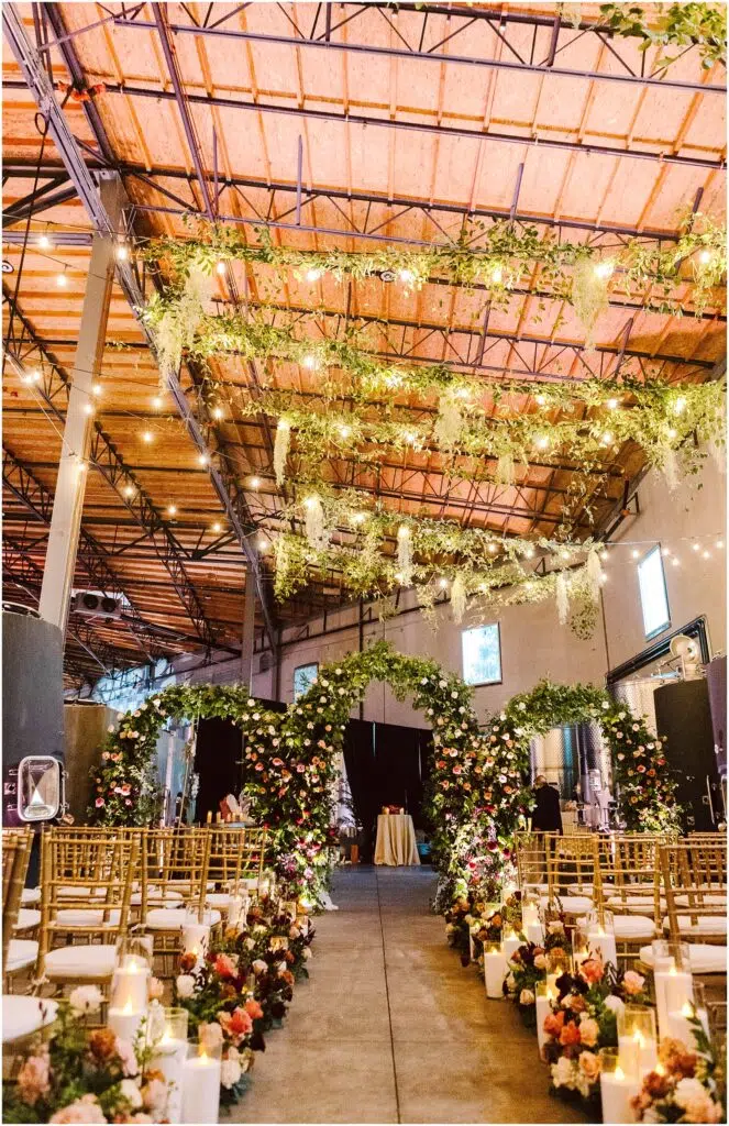 Snohomish Wedding Photographer GSquared Weddings A beautifully decorated indoor wedding aisle with two floral arches adorned with greenery and colorful flowers. Gold chairs line the aisle, and candles in glass holders are placed on the floor. Hanging greenery decorates the ceiling, enhancing the warm ambiance. Serving Seattle, Snohomish and Orlando