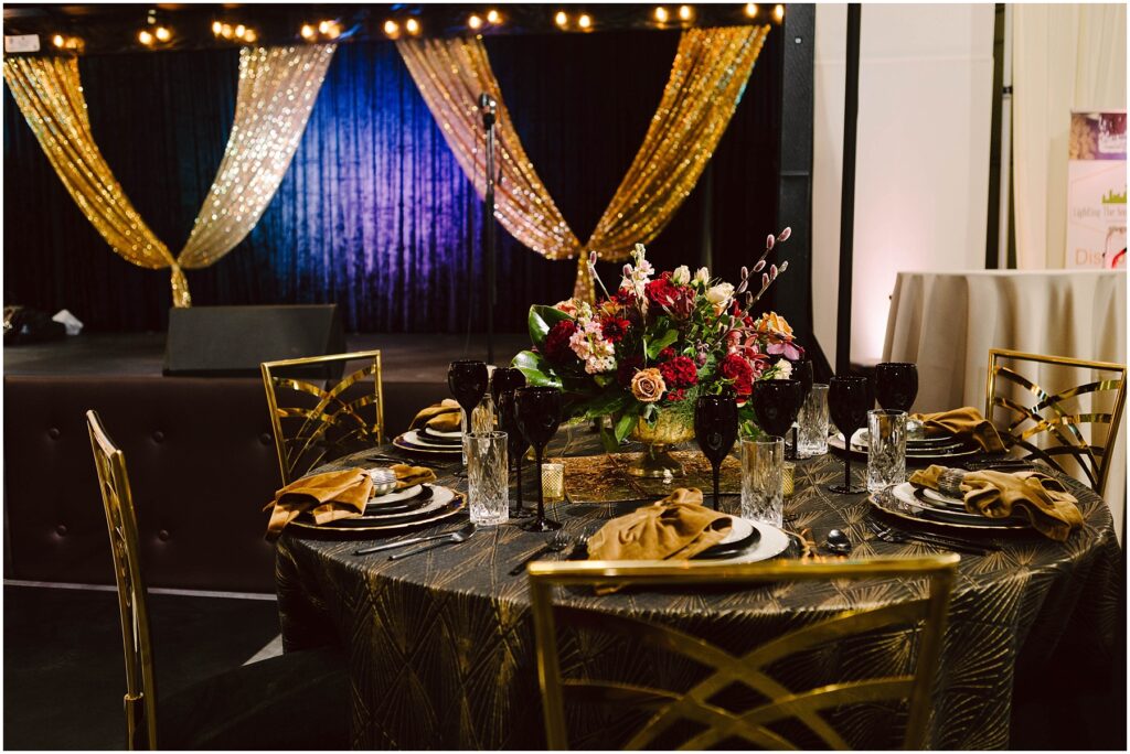 Snohomish Wedding Photographer GSquared Weddings Elegant event table setting with gold and black theme. Features gold chairs, a patterned tablecloth, black and gold dinnerware, and a centerpiece of vibrant flowers. In the background, a stage with gold drapes and string lights. Serving Seattle, Snohomish and Orlando
