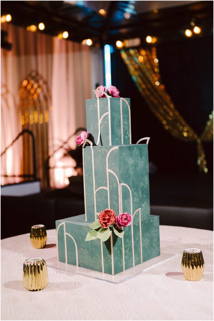 Snohomish Wedding Photographer GSquared Weddings A three-tiered green wedding cake with white geometric patterns, adorned with pink roses on top. It sits on a table with a white textured cloth, surrounded by gold candle holders. The background features draped curtains and decorative golden arches. Serving Seattle, Snohomish and Orlando