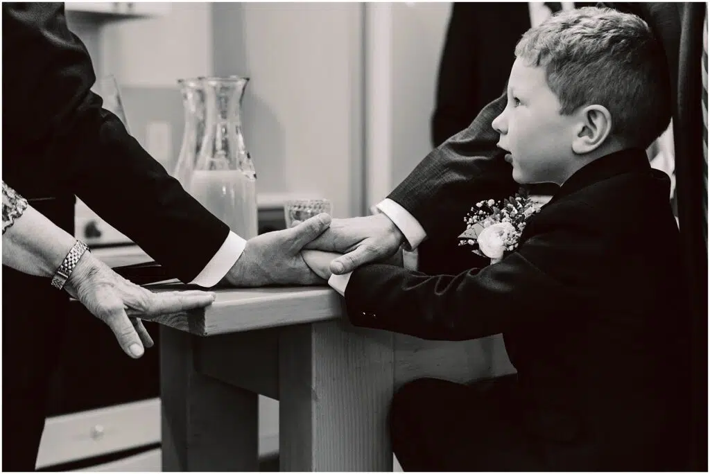 Snohomish Wedding Photographer GSquared Weddings A black and white photo of a young boy in a suit sitting at a table. Several adult hands are placed on top of each other next to him, symbolizing unity or support. A glass pitcher is visible in the background. Serving Seattle, Snohomish and Orlando