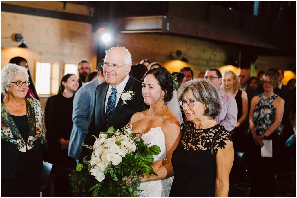 Snohomish Wedding Photographer GSquared Weddings A bride in a white dress holding a bouquet of white flowers stands between two older adults, smiling as they walk down an aisle. Guests are seated on both sides, watching the procession in a warmly lit venue. Serving Seattle, Snohomish and Orlando