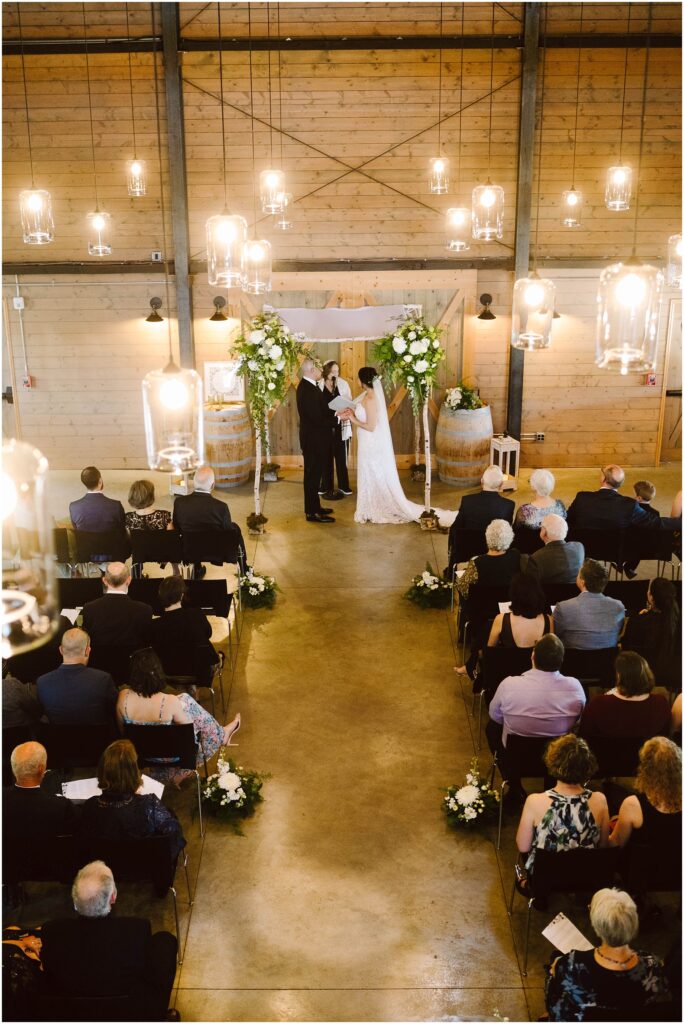 Snohomish Wedding Photographer GSquared Weddings A wedding ceremony in a rustic venue with wooden walls. The couple stands at the altar adorned with flowers and barrels, surrounded by seated guests. Overhead, warm hanging lights illuminate the scene. Serving Seattle, Snohomish and Orlando