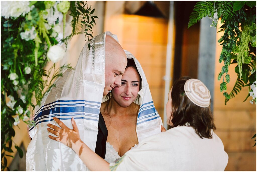 Snohomish Wedding Photographer GSquared Weddings A couple, covered with a white and blue-striped cloth, stands closely together under a canopy. They are joined by a person facing them, wearing a kippah. The setting is adorned with greenery and white flowers, and the atmosphere is serene and intimate. Serving Seattle, Snohomish and Orlando
