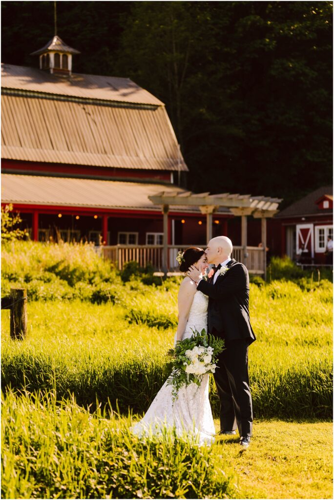 Snohomish Wedding Photographer GSquared Weddings A bride and groom stand in front of a rustic barn surrounded by lush greenery. The bride wears a lace gown and holds a bouquet, while the groom is in a dark suit. They gaze at each other lovingly under the warm sunlight. Serving Seattle, Snohomish and Orlando