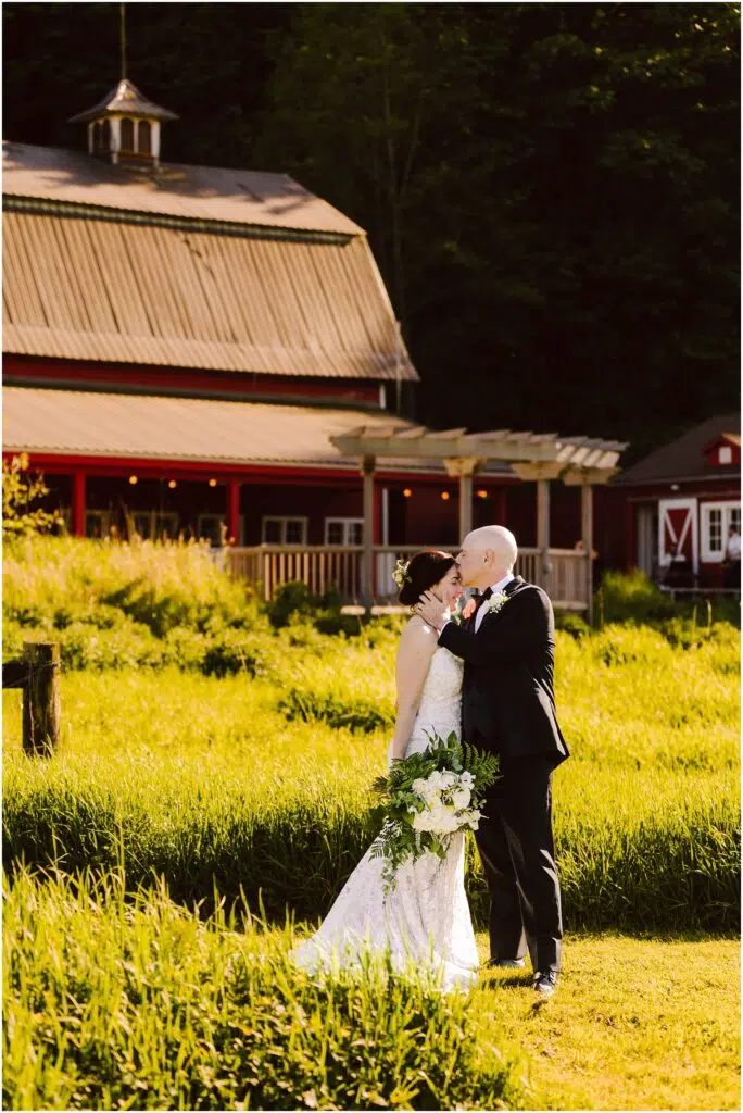Snohomish Wedding Photographer GSquared Weddings A bride and groom stand in front of a rustic barn surrounded by lush greenery. The bride wears a lace gown and holds a bouquet, while the groom is in a dark suit. They gaze at each other lovingly under the warm sunlight. Serving Seattle, Snohomish and Orlando
