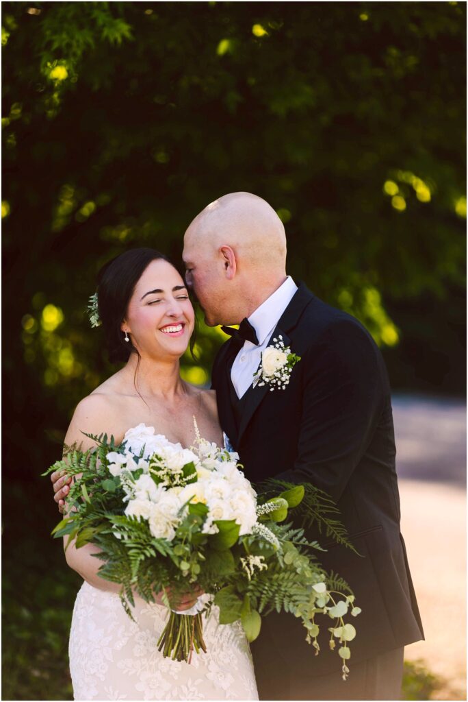 Snohomish Wedding Photographer GSquared Weddings A bride and groom stand outdoors, smiling. The groom, in a black tuxedo, leans in to whisper to the bride, who is wearing a white strapless gown and holding a bouquet of white flowers and greenery. They are surrounded by lush greenery. Serving Seattle, Snohomish and Orlando