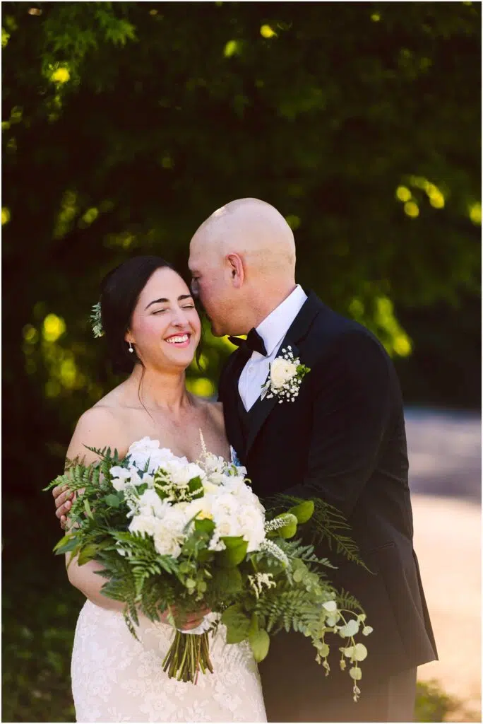 Snohomish Wedding Photographer GSquared Weddings A bride and groom stand outdoors, smiling. The groom, in a black tuxedo, leans in to whisper to the bride, who is wearing a white strapless gown and holding a bouquet of white flowers and greenery. They are surrounded by lush greenery. Serving Seattle, Snohomish and Orlando