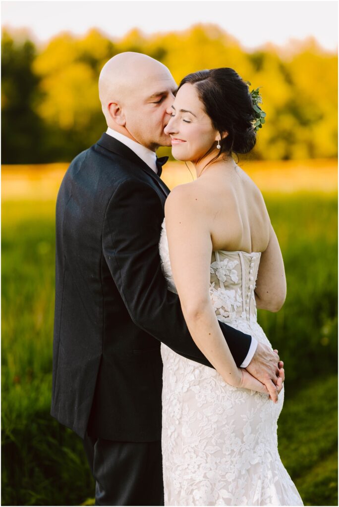 Snohomish Wedding Photographer GSquared Weddings A couple embraces in a grassy field, illuminated by warm sunlight. The man, wearing a black suit, gently kisses the womans face. She is in a white lace strapless dress, smiling with closed eyes. Trees are blurred in the background. Serving Seattle, Snohomish and Orlando