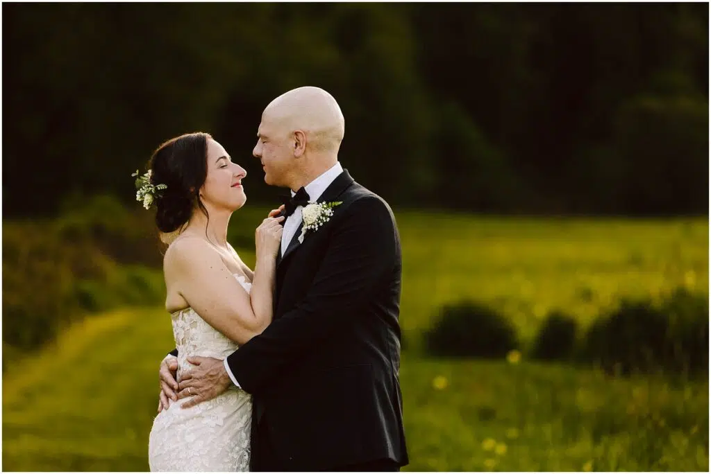 Snohomish Wedding Photographer GSquared Weddings A bride and groom embrace in a field with lush greenery in the background. The bride, in a white lace dress, smiles at the groom, who wears a black suit and bow tie. They are surrounded by a soft, sunlit landscape. Serving Seattle, Snohomish and Orlando