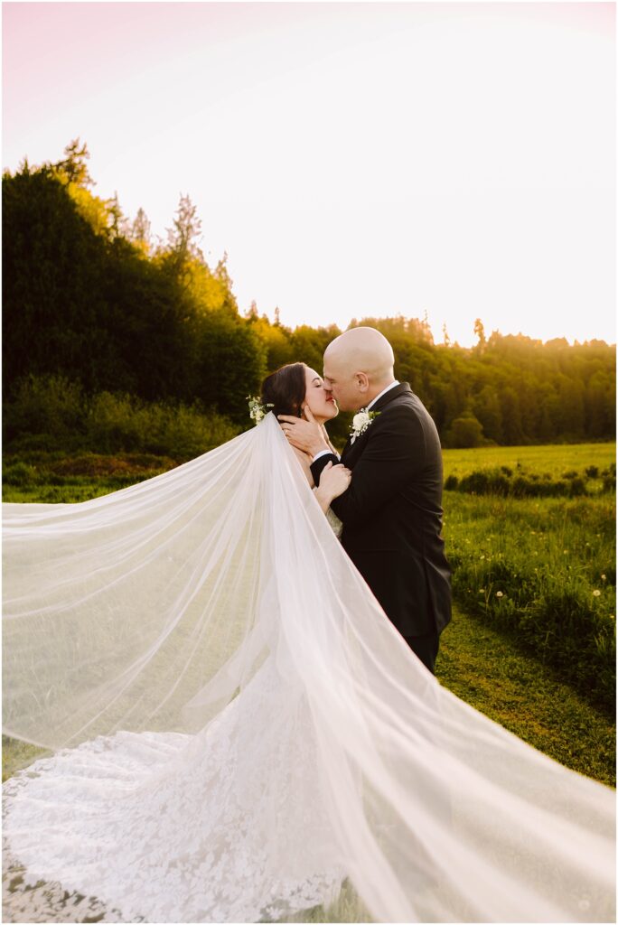 Snohomish Wedding Photographer GSquared Weddings A bride and groom share a kiss in a scenic outdoor setting at sunset. The brides long veil flows dramatically in the breeze, creating a sense of movement. They are standing in a lush green field with trees in the background under a clear sky. Serving Seattle, Snohomish and Orlando