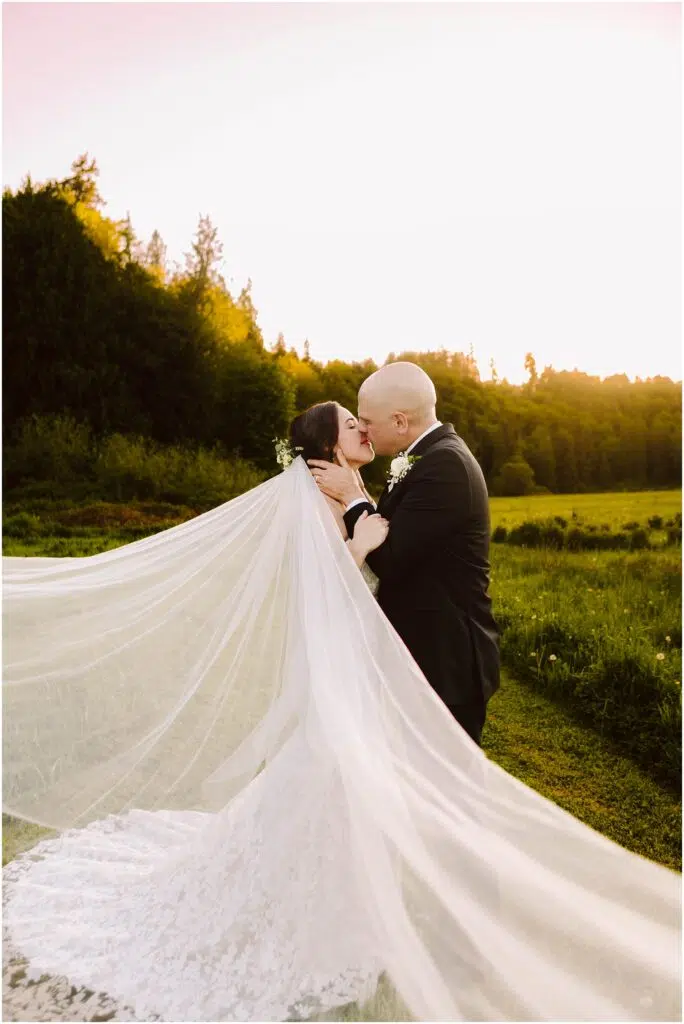 Snohomish Wedding Photographer GSquared Weddings A bride and groom share a kiss in a scenic outdoor setting at sunset. The brides long veil flows dramatically in the breeze, creating a sense of movement. They are standing in a lush green field with trees in the background under a clear sky. Serving Seattle, Snohomish and Orlando