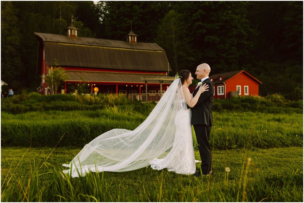 Snohomish Wedding Photographer GSquared Weddings Bride and groom embracing in a grassy field with a red barn in the background. The bride wears a long white gown with a flowing veil, while the groom is in a black suit. The barn has warm lights and a small red building nearby. Serving Seattle, Snohomish and Orlando