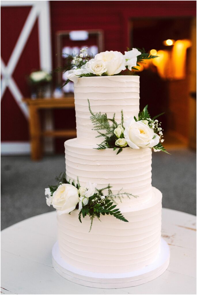 Snohomish Wedding Photographer GSquared Weddings Three-tiered white wedding cake adorned with white roses and green foliage. The cake features a ribbed texture and is displayed indoors, with a red and white wooden wall in the background. Serving Seattle, Snohomish and Orlando