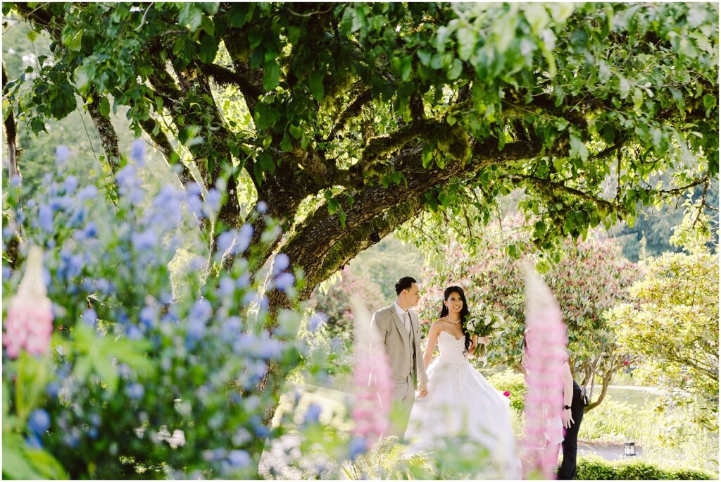 Snohomish Wedding Photographer GSquared Weddings A bride in a white wedding dress and a groom in a light-colored suit walk hand in hand under a large tree. They are surrounded by vibrant flowers and lush greenery, creating a romantic and picturesque garden setting. Serving Seattle, Snohomish and Orlando
