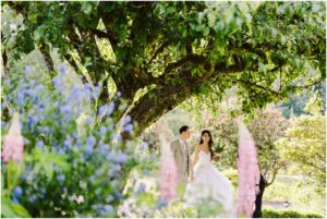 Snohomish Wedding Photographer GSquared Weddings A bride in a white wedding dress and a groom in a light-colored suit walk hand in hand under a large tree. They are surrounded by vibrant flowers and lush greenery, creating a romantic and picturesque garden setting. Serving Seattle, Snohomish and Orlando