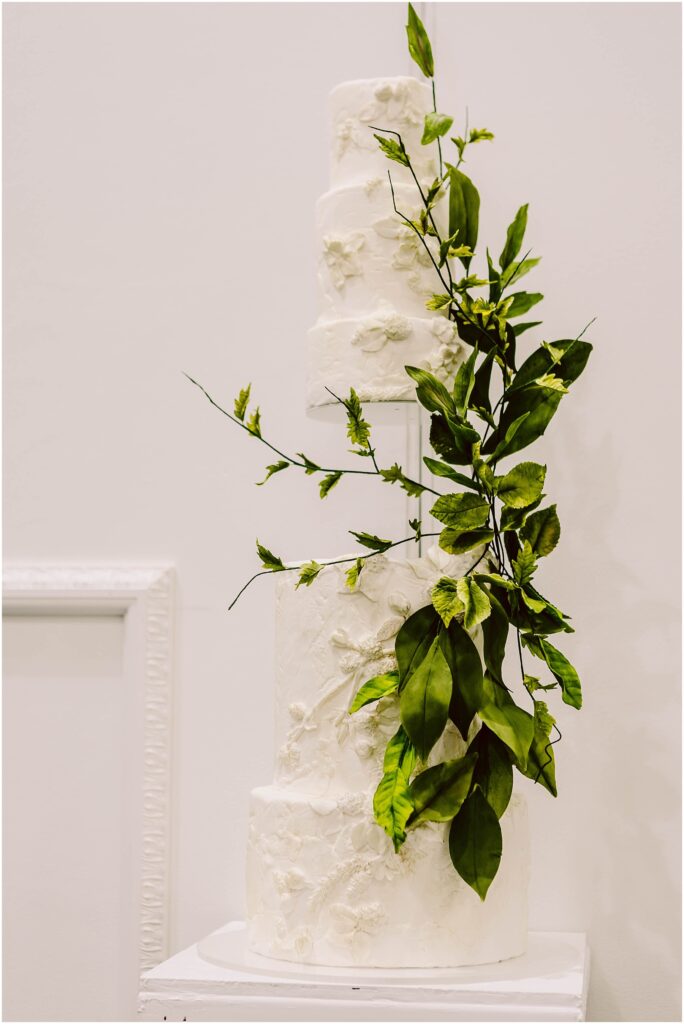 Snohomish Wedding Photographer GSquared Weddings A three-tiered white wedding cake decorated with textured floral patterns and adorned with cascading green leaves, displayed on a white pedestal against a plain background. Serving Seattle, Snohomish and Orlando