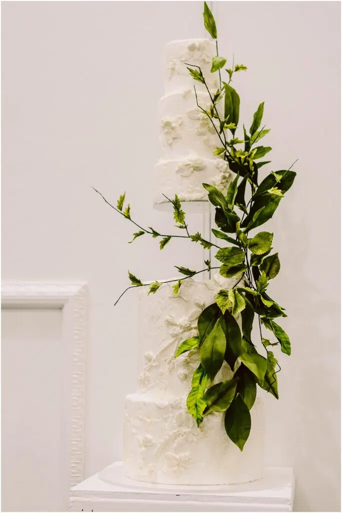 Snohomish Wedding Photographer GSquared Weddings A three-tiered white wedding cake decorated with textured floral patterns and adorned with cascading green leaves, displayed on a white pedestal against a plain background. Serving Seattle, Snohomish and Orlando