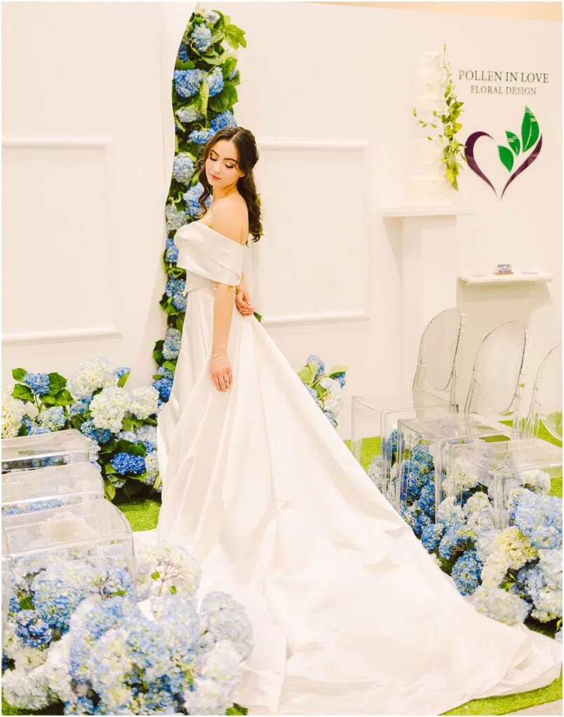 Snohomish Wedding Photographer GSquared Weddings A woman in an elegant off-the-shoulder white gown poses beside a display of blue and white floral arrangements. The setting includes clear chairs and a sign that reads Pollen in Love Floral Design with a heart-shaped logo. Serving Seattle, Snohomish and Orlando
