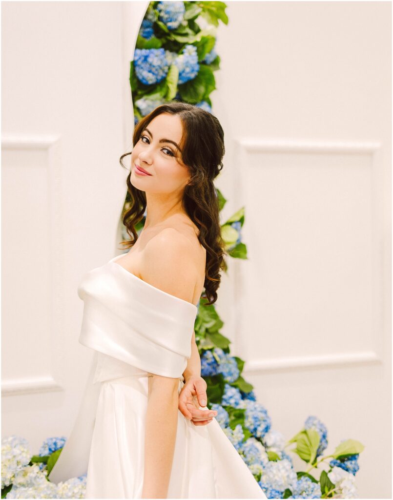 Snohomish Wedding Photographer GSquared Weddings A woman in an elegant off-shoulder white dress stands gracefully in front of a backdrop decorated with blue and white hydrangeas. She has long, wavy brown hair and is smiling softly, creating a serene and joyful atmosphere. Serving Seattle, Snohomish and Orlando