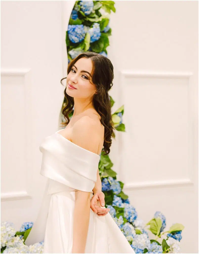 Snohomish Wedding Photographer GSquared Weddings A woman in an elegant off-shoulder white dress stands gracefully in front of a backdrop decorated with blue and white hydrangeas. She has long, wavy brown hair and is smiling softly, creating a serene and joyful atmosphere. Serving Seattle, Snohomish and Orlando