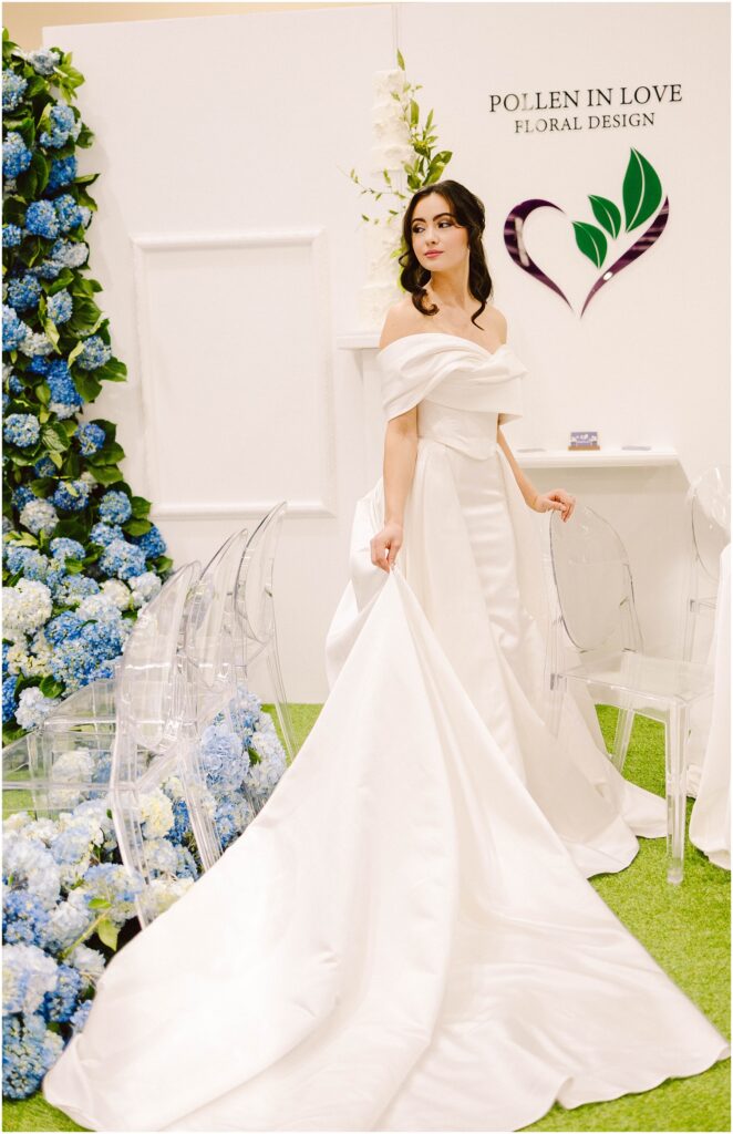 Snohomish Wedding Photographer GSquared Weddings Bride in an elegant white gown with a long train stands in front of a floral display featuring blue and white flowers. Clear chairs line the area. A heart-shaped floral design and the words Pollen in Love Floral Design are on the wall. Serving Seattle, Snohomish and Orlando