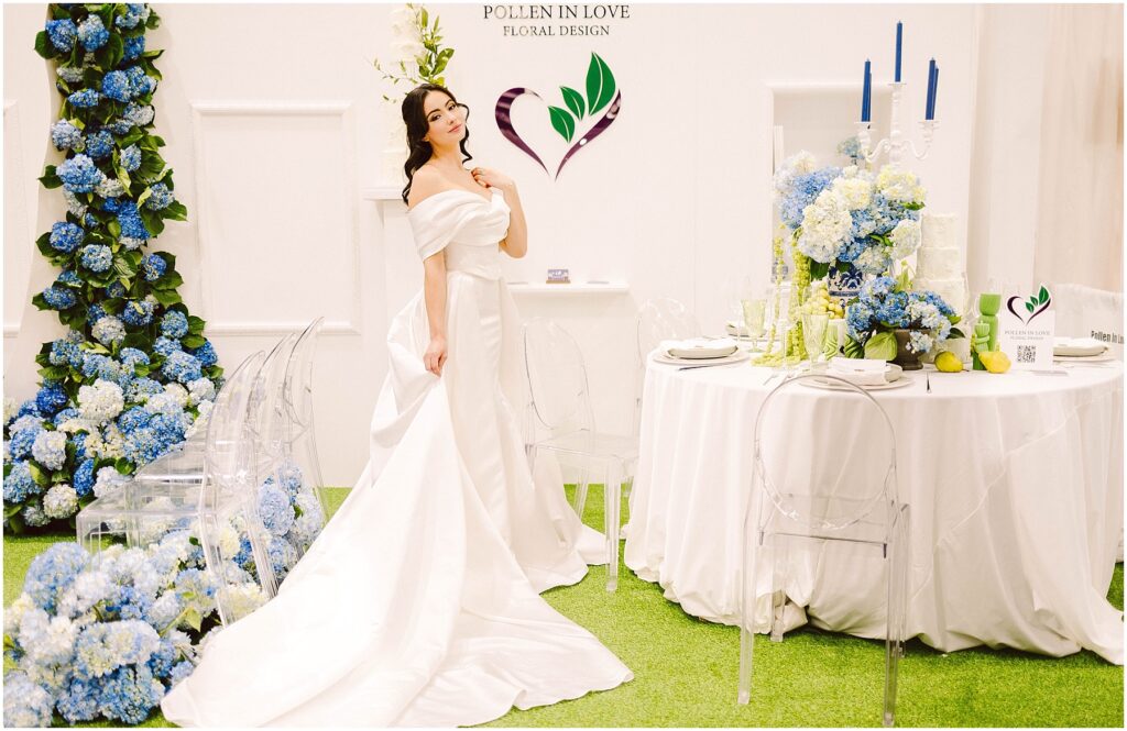 Snohomish Wedding Photographer GSquared Weddings A woman in an elegant white gown stands in a floral-themed setup, next to a white round table with blue floral and candle arrangements. Pollen in Love Floral Design is displayed on the wall. Clear chairs surround the table on a grass-like carpet. Serving Seattle, Snohomish and Orlando