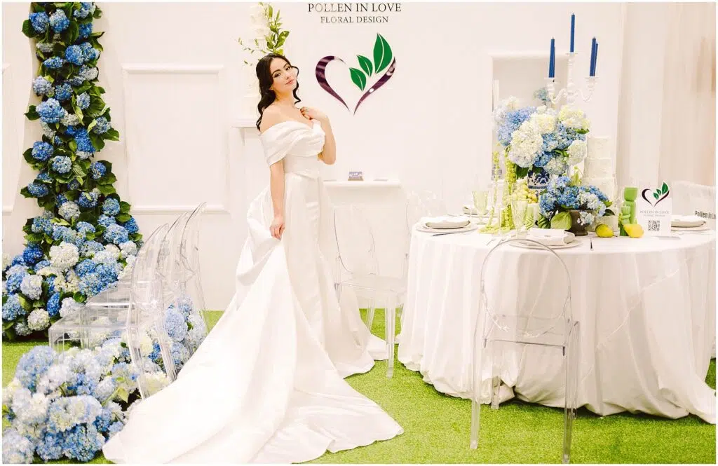 Snohomish Wedding Photographer GSquared Weddings A woman in an elegant white gown stands in a floral-themed setup, next to a white round table with blue floral and candle arrangements. Pollen in Love Floral Design is displayed on the wall. Clear chairs surround the table on a grass-like carpet. Serving Seattle, Snohomish and Orlando