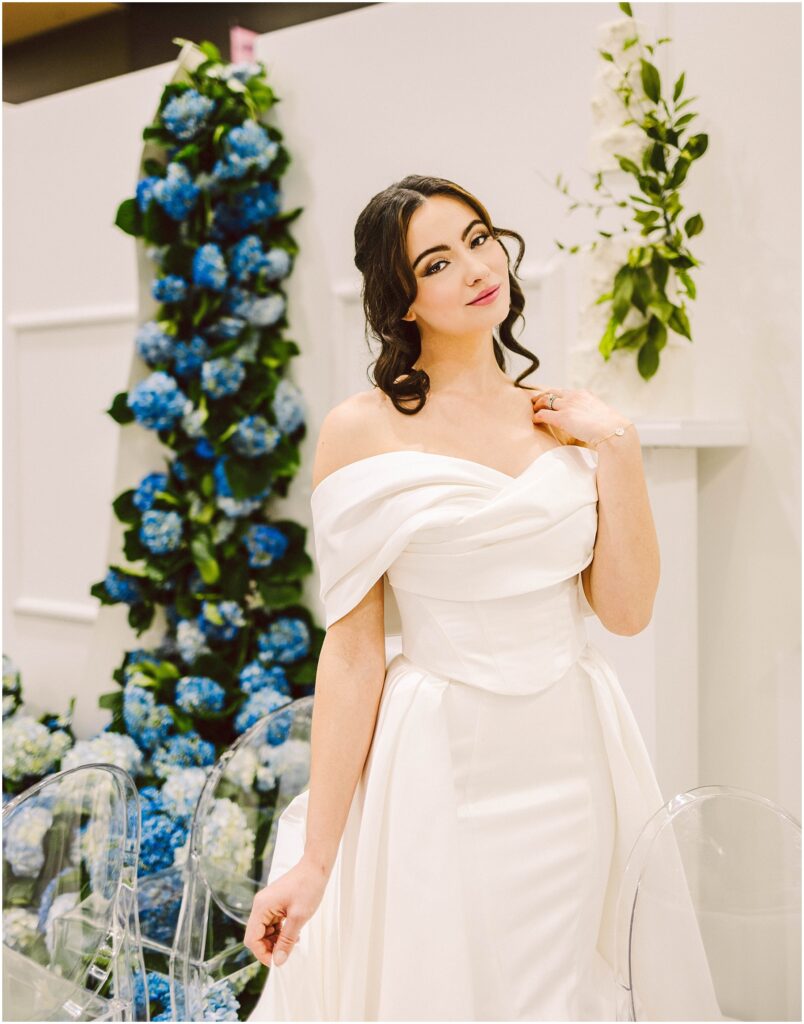 Snohomish Wedding Photographer GSquared Weddings A woman in an elegant off-shoulder white dress stands gracefully beside a tall floral arrangement of blue and white flowers. She is in a well-lit setting with clear chairs visible nearby, exuding a serene and poised demeanor. Serving Seattle, Snohomish and Orlando