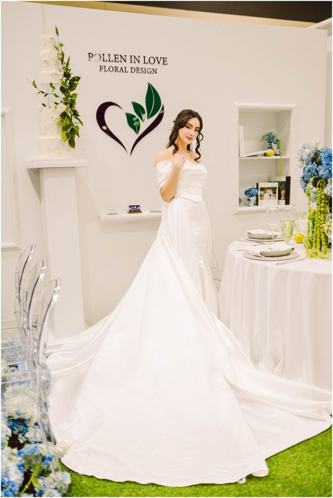 Snohomish Wedding Photographer GSquared Weddings A woman in a white wedding dress poses in front of a Pollen in Love Floral Design sign. The setting includes a table with blue flowers, elegant tableware, and a long train extending from the dress. The space has a sophisticated floral theme. Serving Seattle, Snohomish and Orlando