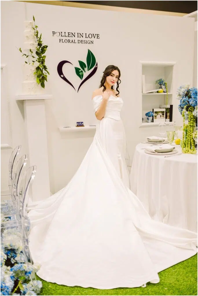 Snohomish Wedding Photographer GSquared Weddings A woman in a white wedding dress poses in front of a Pollen in Love Floral Design sign. The setting includes a table with blue flowers, elegant tableware, and a long train extending from the dress. The space has a sophisticated floral theme. Serving Seattle, Snohomish and Orlando