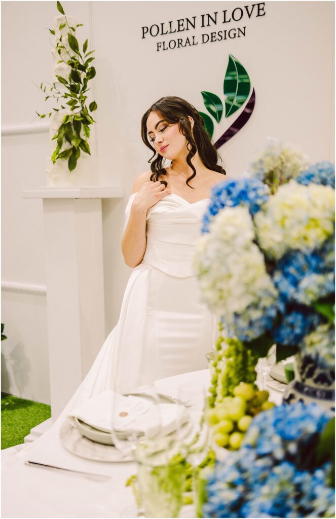 Snohomish Wedding Photographer GSquared Weddings A woman in an elegant white dress stands beside a table with blue and green floral arrangements. She looks down thoughtfully, with her hand near her shoulder. The backdrop features the words Pollen in Love Floral Design and leafy decorations. Serving Seattle, Snohomish and Orlando