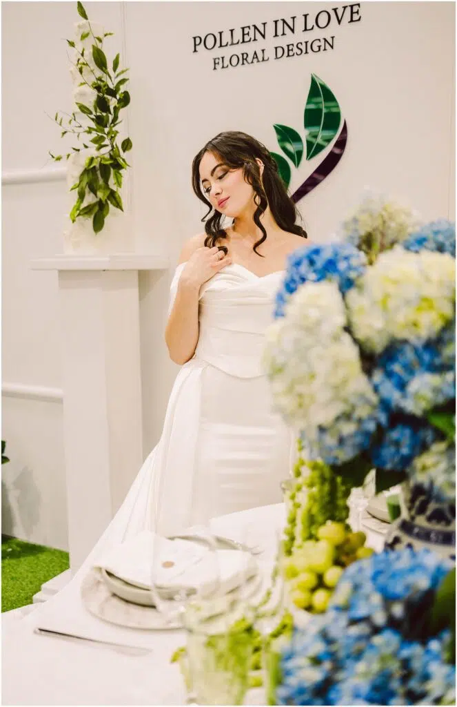 Snohomish Wedding Photographer GSquared Weddings A woman in an elegant white dress stands beside a table with blue and green floral arrangements. She looks down thoughtfully, with her hand near her shoulder. The backdrop features the words Pollen in Love Floral Design and leafy decorations. Serving Seattle, Snohomish and Orlando