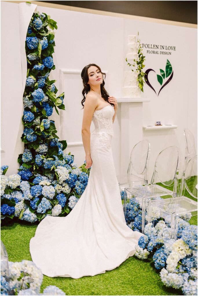 Snohomish Wedding Photographer GSquared Weddings A bride in a strapless white wedding dress poses gracefully beside floral arrangements of blue and white hydrangeas. Clear chairs and a floral design logo are in the background. Serving Seattle, Snohomish and Orlando