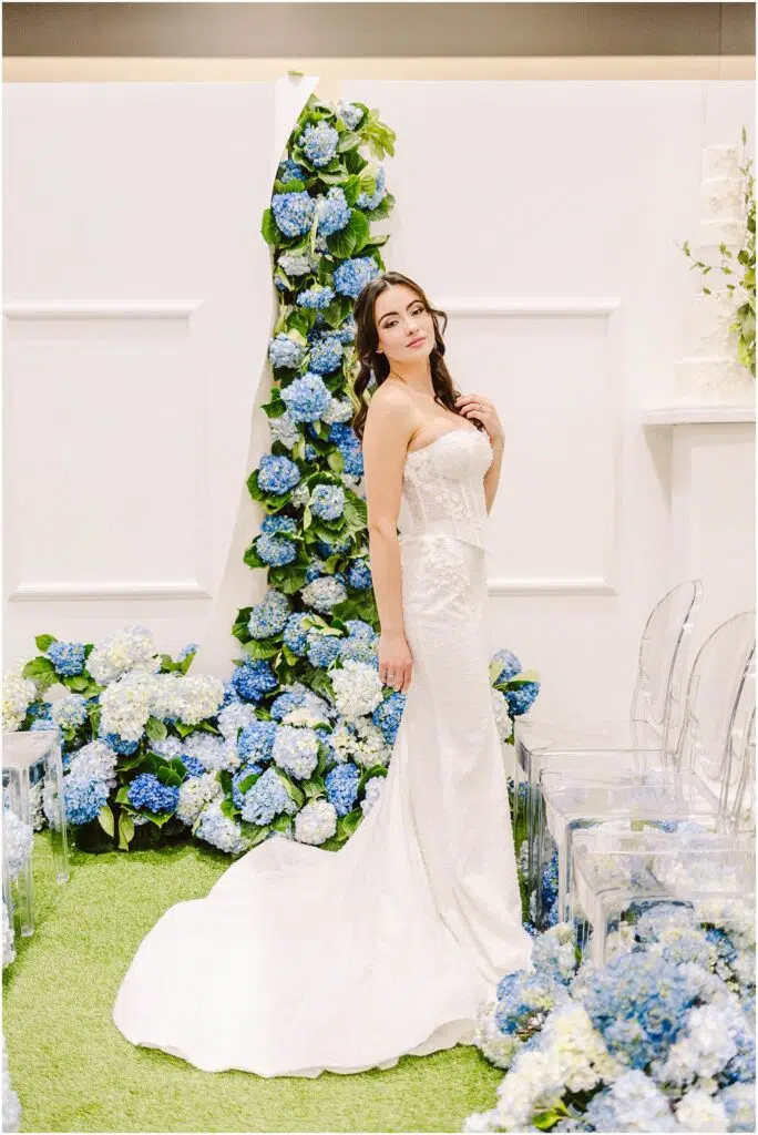 Snohomish Wedding Photographer GSquared Weddings A bride in a strapless white gown stands by a floral arch adorned with blue and white hydrangeas. She poses on green grass next to clear chairs, with her hair styled in loose waves. The setting is elegant and bright. Serving Seattle, Snohomish and Orlando