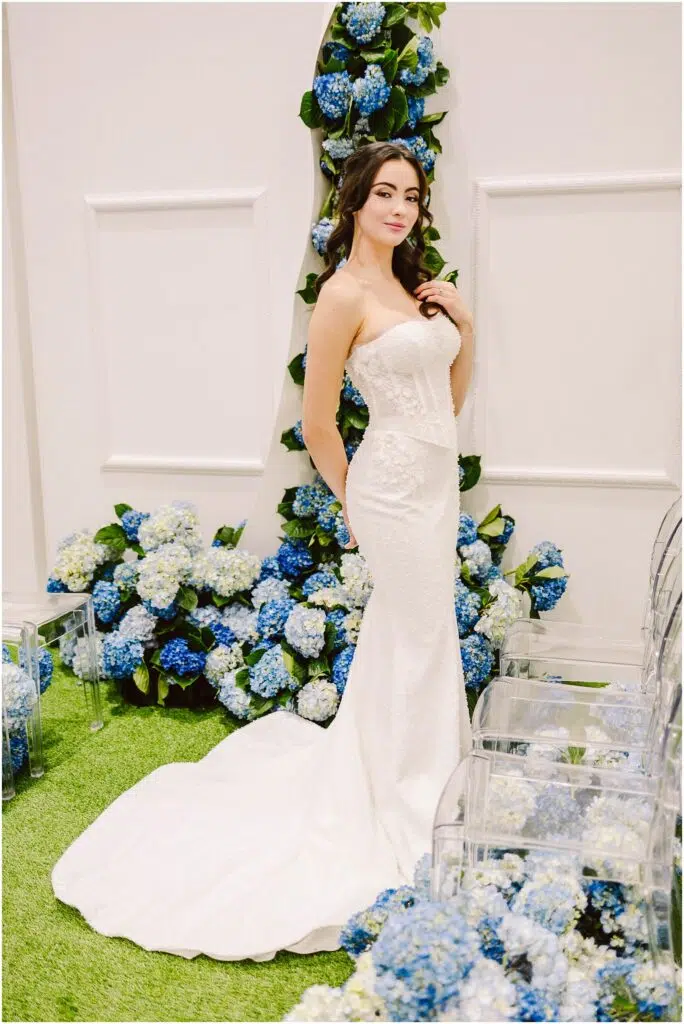 Snohomish Wedding Photographer GSquared Weddings A woman in a strapless white wedding dress stands on green grass. She is surrounded by blue and white hydrangea flowers. Clear chairs are visible on the side. The setting is elegant and floral. Serving Seattle, Snohomish and Orlando