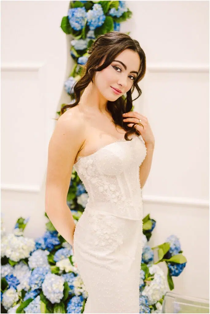 Snohomish Wedding Photographer GSquared Weddings A woman in a strapless white wedding dress stands in front of a backdrop adorned with blue and white flowers. She has long, wavy hair and is posing with one hand on her hip, smiling softly at the camera. Serving Seattle, Snohomish and Orlando