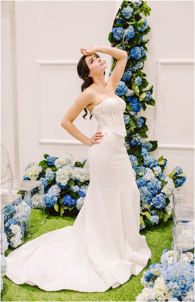 Snohomish Wedding Photographer GSquared Weddings A woman in a strapless white wedding dress poses with one hand on her hip and the other on her forehead. She stands on grass surrounded by blue and white flowers. Transparent chairs are visible nearby. Serving Seattle, Snohomish and Orlando