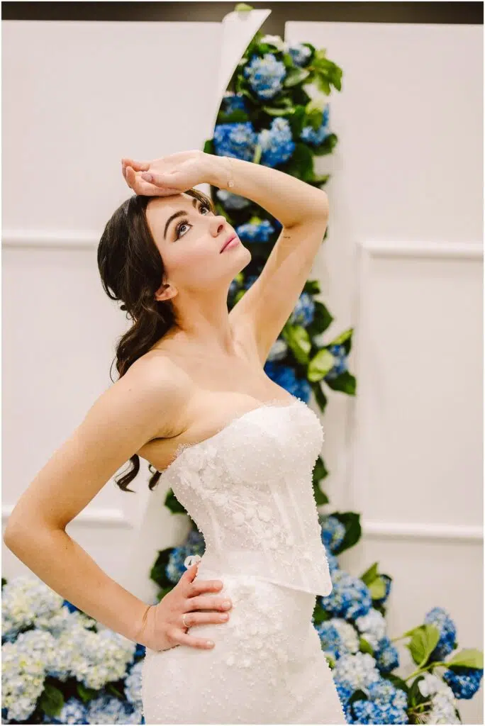 Snohomish Wedding Photographer GSquared Weddings A bride in a strapless, white lace wedding dress poses dramatically. Her hand rests on her forehead, and she stands in front of a backdrop adorned with blue and white flowers. Her dark hair is styled elegantly. Serving Seattle, Snohomish and Orlando