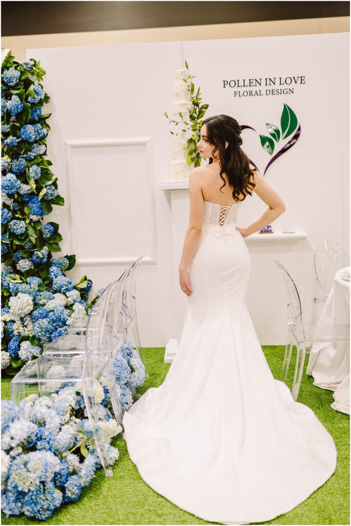 Snohomish Wedding Photographer GSquared Weddings A bride in a strapless white gown stands with her back to the camera, in front of a display with blue hydrangeas and a white floral-themed backdrop. The setting includes transparent chairs and a sign reading Pollen in Love Floral Design. Serving Seattle, Snohomish and Orlando