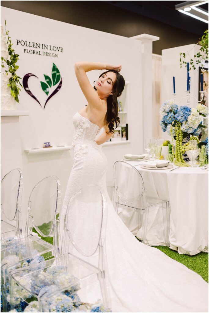 Snohomish Wedding Photographer GSquared Weddings A woman in a strapless white wedding gown poses with her arm raised, standing next to a table with a white tablecloth and blue flowers. Transparent chairs surround the table. The backdrop features floral designs and the words Pollen In Love Floral Design. Serving Seattle, Snohomish and Orlando