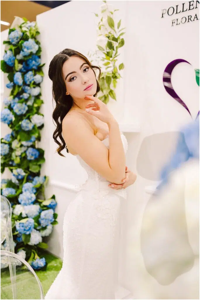 Snohomish Wedding Photographer GSquared Weddings A woman in a white strapless dress stands in a floral-themed setting with blue and green hydrangeas. She is gazing at the camera with one hand touching her face. The background features floral decor and a partial logo. Serving Seattle, Snohomish and Orlando